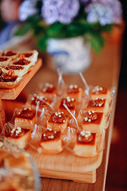 A variety of desserts arranged neatly on wooden trays, featuring caramel-topped squares with small white decorations. In the background, a blurred floral arrangement in a decorative vase adds to the setting's elegance.