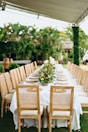 A long table covered with a white tablecloth is elegantly set for a banquet. The table is lined with ornate wooden chairs featuring wicker backs and beige cushions. A lush floral arrangement runs along the center of the table, adding a touch of nature and sophistication. The background is filled with green foliage, suggesting an outdoor setting under a canopy.