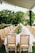 A long table covered with a white tablecloth is elegantly set for a banquet. The table is lined with ornate wooden chairs featuring wicker backs and beige cushions. A lush floral arrangement runs along the center of the table, adding a touch of nature and sophistication. The background is filled with green foliage, suggesting an outdoor setting under a canopy.