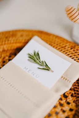 A well-arranged table setting featuring a folded beige napkin with a menu card tucked inside. The menu card has an image of a sprig of rosemary and the word 'Menú' printed on it. The background includes a woven, circular placemat and a honey dipper.