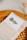 A well-arranged table setting featuring a folded beige napkin with a menu card tucked inside. The menu card has an image of a sprig of rosemary and the word 'Menú' printed on it. The background includes a woven, circular placemat and a honey dipper.