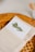 A well-arranged table setting featuring a folded beige napkin with a menu card tucked inside. The menu card has an image of a sprig of rosemary and the word 'Menú' printed on it. The background includes a woven, circular placemat and a honey dipper.