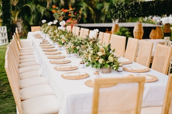 A long outdoor dining table is elegantly set with beige cushioned chairs and a pristine white tablecloth. Placed along the center are lush floral arrangements featuring greenery and white flowers. Each setting has a round, woven placemat and a small vase. The background includes greenery and decorative elements such as hanging lanterns and structures.