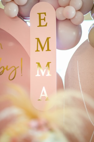 A pink vertical sign with gold lettering spelling 'Emma' is surrounded by clusters of pale pink and white balloons. Additional decorative elements include a faintly visible gold script in the background.
