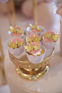 Cupcakes are elegantly displayed on a decorative gold stand. Each cupcake is adorned with intricate gold butterfly decorations and pink icing. The setup creates a luxurious and delicate look.