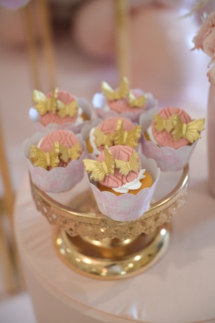 Cupcakes are elegantly displayed on a decorative gold stand. Each cupcake is adorned with intricate gold butterfly decorations and pink icing. The setup creates a luxurious and delicate look.
