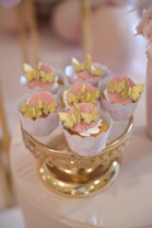 Cupcakes are elegantly displayed on a decorative gold stand. Each cupcake is adorned with intricate gold butterfly decorations and pink icing. The setup creates a luxurious and delicate look.