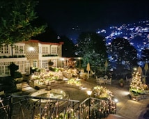 A beautifully lit courtyard at night features a charming stone building adorned with festive lights. The foreground includes a small fountain and decorative plants, while the patio area is set with tables and chairs. Across the landscape, trees are seen dimly, as the background reveals a softly glowing cityscape.