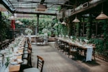 A beautifully set dining area with long tables adorned with elegant tableware and floral arrangements. The space is enclosed by glass walls, allowing natural light to illuminate the room. The walls are covered in lush greenery, creating a cozy and inviting atmosphere.
