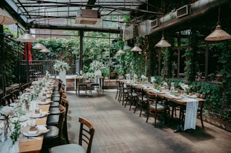 A beautifully set dining area with long tables adorned with elegant tableware and floral arrangements. The space is enclosed by glass walls, allowing natural light to illuminate the room. The walls are covered in lush greenery, creating a cozy and inviting atmosphere.