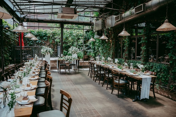 A beautifully set dining area with long tables adorned with elegant tableware and floral arrangements. The space is enclosed by glass walls, allowing natural light to illuminate the room. The walls are covered in lush greenery, creating a cozy and inviting atmosphere.