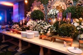An elegant buffet setup decorated with lush green foliage and white flowers, accented by ornate gold elements. A large mirror with intricate designs is partially visible, surrounded by candles in tall glass holders. Stacks of white plates and serving utensils are neatly arranged on a wooden table, ready for use.