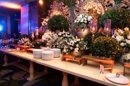 An elegant buffet setup decorated with lush green foliage and white flowers, accented by ornate gold elements. A large mirror with intricate designs is partially visible, surrounded by candles in tall glass holders. Stacks of white plates and serving utensils are neatly arranged on a wooden table, ready for use.