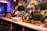 An elegant buffet setup decorated with lush green foliage and white flowers, accented by ornate gold elements. A large mirror with intricate designs is partially visible, surrounded by candles in tall glass holders. Stacks of white plates and serving utensils are neatly arranged on a wooden table, ready for use.