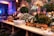 An elegant buffet setup decorated with lush green foliage and white flowers, accented by ornate gold elements. A large mirror with intricate designs is partially visible, surrounded by candles in tall glass holders. Stacks of white plates and serving utensils are neatly arranged on a wooden table, ready for use.