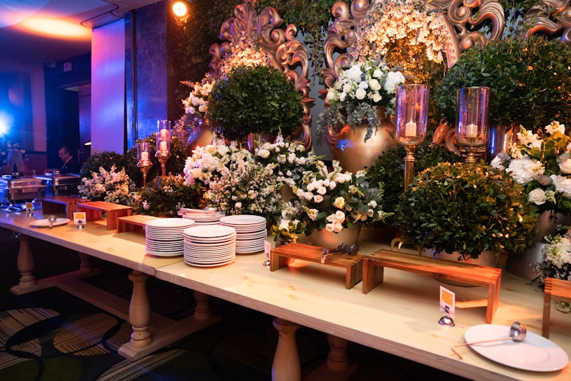 An elegant buffet setup decorated with lush green foliage and white flowers, accented by ornate gold elements. A large mirror with intricate designs is partially visible, surrounded by candles in tall glass holders. Stacks of white plates and serving utensils are neatly arranged on a wooden table, ready for use.