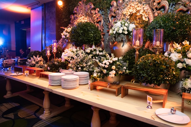 An elegant buffet setup decorated with lush green foliage and white flowers, accented by ornate gold elements. A large mirror with intricate designs is partially visible, surrounded by candles in tall glass holders. Stacks of white plates and serving utensils are neatly arranged on a wooden table, ready for use.