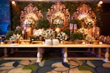 An elaborate and ornate buffet setup featuring a long wooden table adorned with dishes and serving utensils. Behind the table, large decorative golden floral carvings are accentuated by green foliage and white floral arrangements. Ornate candelabras and decorative vases add to the luxurious setup, while the patterned carpet below complements the elegant display.