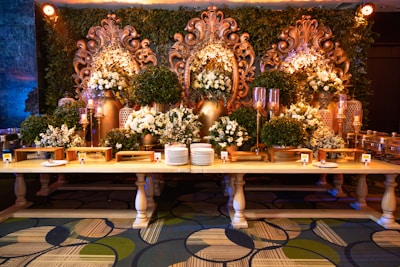 An elaborate and ornate buffet setup featuring a long wooden table adorned with dishes and serving utensils. Behind the table, large decorative golden floral carvings are accentuated by green foliage and white floral arrangements. Ornate candelabras and decorative vases add to the luxurious setup, while the patterned carpet below complements the elegant display.