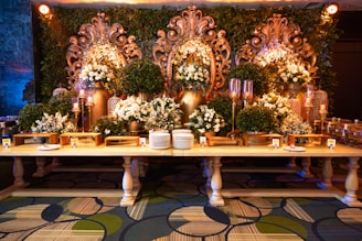 An elaborate and ornate buffet setup featuring a long wooden table adorned with dishes and serving utensils. Behind the table, large decorative golden floral carvings are accentuated by green foliage and white floral arrangements. Ornate candelabras and decorative vases add to the luxurious setup, while the patterned carpet below complements the elegant display.