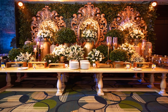 An elaborate and ornate buffet setup featuring a long wooden table adorned with dishes and serving utensils. Behind the table, large decorative golden floral carvings are accentuated by green foliage and white floral arrangements. Ornate candelabras and decorative vases add to the luxurious setup, while the patterned carpet below complements the elegant display.