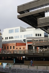 A modern urban scene features an architectural blend of traditional brickwork and contemporary design. A cyclist rides alongside a riverfront, while banners in various colors adorn the railing. Large windows and structured metal elements contrast with the warm brick tones of the older building.