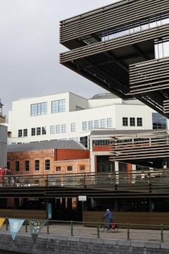 A modern urban scene features an architectural blend of traditional brickwork and contemporary design. A cyclist rides alongside a riverfront, while banners in various colors adorn the railing. Large windows and structured metal elements contrast with the warm brick tones of the older building.