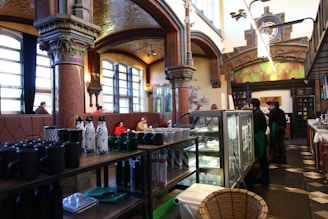 A spacious, vintage-style cafe interior featuring ornate columns and high ceilings. Large windows allow natural light to illuminate the room, where several patrons are seated in leather booths. Shelves stocked with cups and bottles are in the foreground, while staff in green aprons work behind a glass counter that displays menu items.