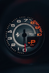 A close-up of a car's tachometer displaying RPM measurements with a red needle pointing at zero. It features white and orange numbering on a dark gray background, with an orange 'P auto' indicator symbol prominent in the lower right.