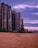 A row of tall modern apartment buildings line a sandy beach under a sky filled with purple and pink hues from the setting or rising sun. The beach is deserted, creating an atmosphere of calm and solitude.