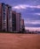 A row of tall modern apartment buildings line a sandy beach under a sky filled with purple and pink hues from the setting or rising sun. The beach is deserted, creating an atmosphere of calm and solitude.