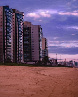 A row of tall modern apartment buildings line a sandy beach under a sky filled with purple and pink hues from the setting or rising sun. The beach is deserted, creating an atmosphere of calm and solitude.