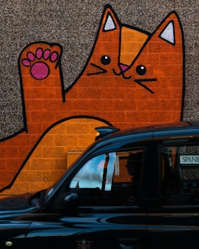 A large, colorful mural of an orange cartoon cat with a playful expression is painted on a brick wall. The cat's paw is raised, with pink paw pads visible. In the foreground, a black taxi cab passes by, partially obscuring the lower part of the mural.
