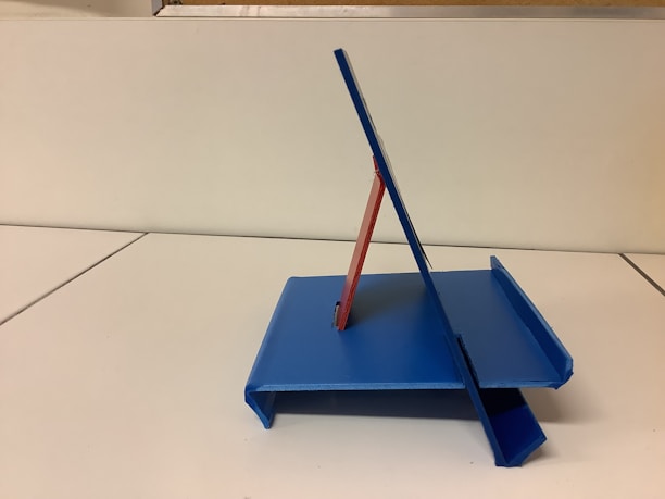 A blue and red plastic or metal stand is positioned on a white surface. The structure has an angular design and appears to be purposefully crafted for holding or supporting objects, possibly books or electronic devices. The background is plain and unobtrusive.