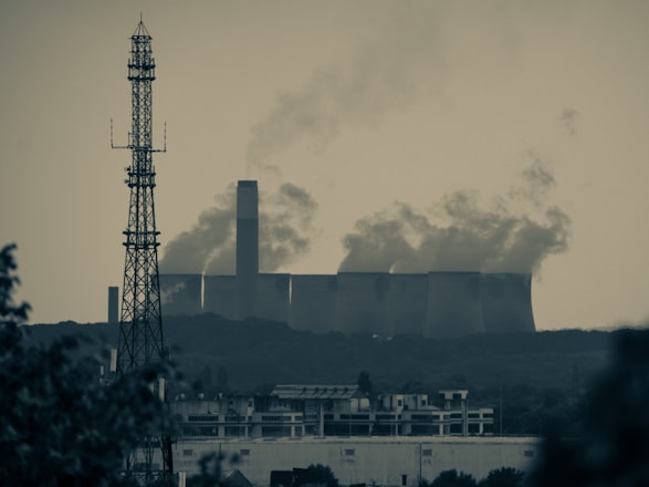 A large industrial facility emits substantial amounts of smoke from multiple tall chimneys, set against a hazy sky. In the foreground, a tall metal radio or cell tower rises above a sprawling urban landscape, featuring residential or commercial buildings. The scene conveys a sense of urban-industrial juxtaposition amidst environmental concerns.