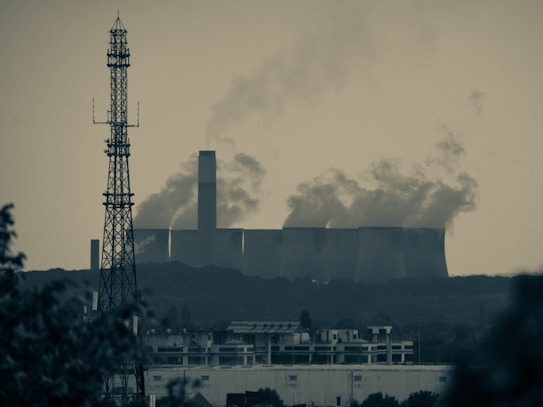 A large industrial facility emits substantial amounts of smoke from multiple tall chimneys, set against a hazy sky. In the foreground, a tall metal radio or cell tower rises above a sprawling urban landscape, featuring residential or commercial buildings. The scene conveys a sense of urban-industrial juxtaposition amidst environmental concerns.