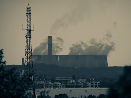 A large industrial facility emits substantial amounts of smoke from multiple tall chimneys, set against a hazy sky. In the foreground, a tall metal radio or cell tower rises above a sprawling urban landscape, featuring residential or commercial buildings. The scene conveys a sense of urban-industrial juxtaposition amidst environmental concerns.