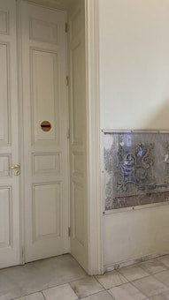 A light-colored wooden door with ornate paneling and gold handles is situated on the left. There is a small circular sign with red and yellow horizontal stripes on one panel. To the right of the door, a piece of abstract art or a textured metal panel is mounted on the wall above a tiled floor.