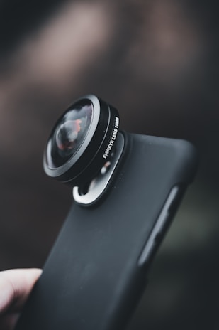 A close-up view of a smartphone with an attached fisheye lens, featuring a smooth, dark protective case. The background is softly blurred, emphasizing the focus on the lens attachment.