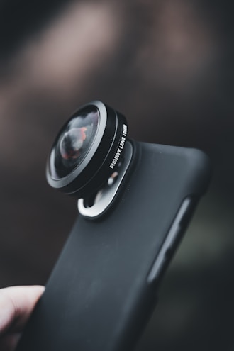 A close-up view of a smartphone with an attached fisheye lens, featuring a smooth, dark protective case. The background is softly blurred, emphasizing the focus on the lens attachment.