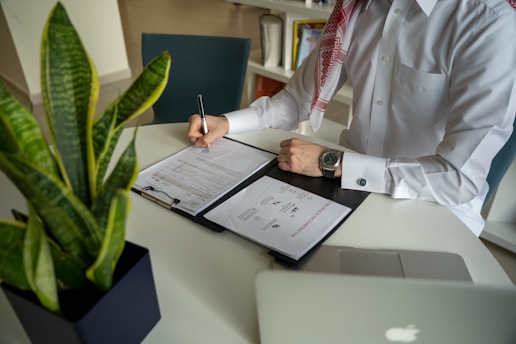 A person wearing a white shirt is sitting at a desk, writing on a document inside a black binder. A watch can be seen on their wrist. A green potted plant is on the desk, along with a laptop. Shelves and books are visible in the background.