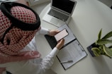 A person wearing a red and white keffiyeh and white clothing is seated at a round table, holding a smartphone. In front of them is an open laptop and a document placed inside a folder with a pen resting on top. A potted plant is also on the table, indicating a business or office setting.