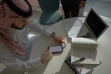 A person wearing traditional Middle Eastern attire is sitting at a table. The table holds a smartphone, an open book, and a laptop, along with some scattered documents. The lighting is natural, coming from a window, partially illuminating the setting.