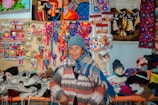 A person wearing a blue knit hat and multicolored poncho stands in front of a vibrant display of handmade crafts and textiles. The backdrop features colorful woven art, textiles, and small souvenirs, showcasing a variety of patterns and bright colors.