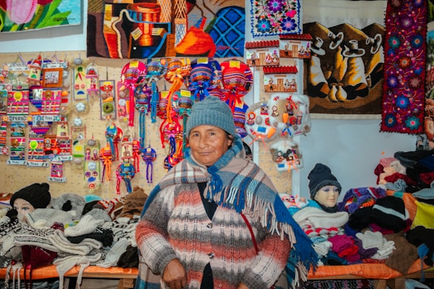 A person wearing a blue knit hat and multicolored poncho stands in front of a vibrant display of handmade crafts and textiles. The backdrop features colorful woven art, textiles, and small souvenirs, showcasing a variety of patterns and bright colors.