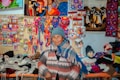 A person wearing a blue knit hat and multicolored poncho stands in front of a vibrant display of handmade crafts and textiles. The backdrop features colorful woven art, textiles, and small souvenirs, showcasing a variety of patterns and bright colors.