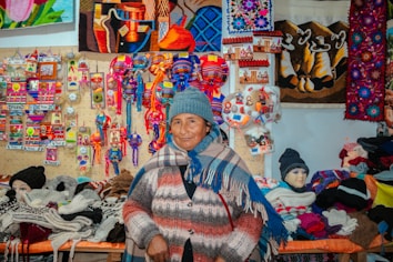 A person wearing a blue knit hat and multicolored poncho stands in front of a vibrant display of handmade crafts and textiles. The backdrop features colorful woven art, textiles, and small souvenirs, showcasing a variety of patterns and bright colors.