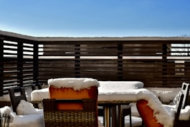 A patio with outdoor furniture, including a table and multiple chairs, is covered in a layer of snow. The patio is enclosed by a wooden fence, and the sky is clear with a bright blue hue.