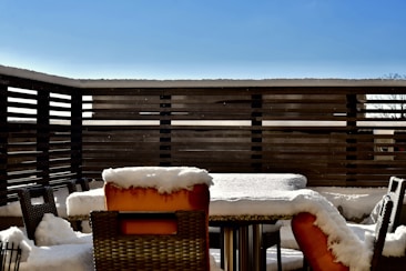 A patio with outdoor furniture, including a table and multiple chairs, is covered in a layer of snow. The patio is enclosed by a wooden fence, and the sky is clear with a bright blue hue.