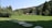 A lush green field is bordered by dense trees and bushes, leading to a cluster of rustic wooden cabins. The setting is backed by hills covered with thick pine forests under a clear sky. Stone pathways cross the vibrant grass, enhancing the idyllic, peaceful atmosphere.